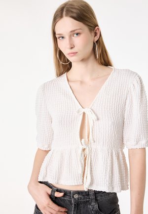 Giovane donna con capelli biondi lisci indossa una blusa bianca texturizzata con nodo sul davanti e maniche a sbuffo, abbinata a jeans neri in denim.