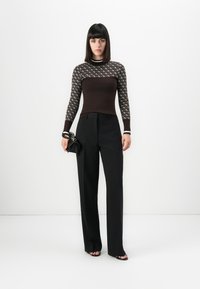 Mujer con cabello negro lacio que lleva una blusa de manga larga con estampado negro y marrón, pantalones de pierna ancha negros y sostiene un pequeño bolso negro.