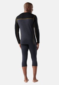 Twee-delig sportonder kleding set bestaande uit een aansluitend zwart langemouwen shirt en donkergrijze capri broeken met gele accentstiksels.