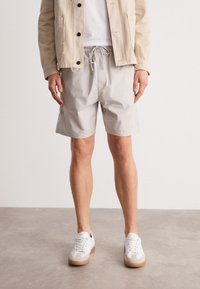 Lichtgrijze shorts met een elastische tailleband en koord, gemaakt van een gladde stof. Gepaard met witte sneakers met lichtbruine zolen.