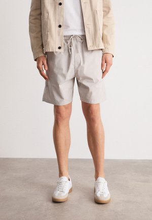 Shorts gris clair avec une ceinture élastique et un cordon de serrage, fabriqués en tissu lisse. Associés à des baskets blanches avec des semelles marron clair.
