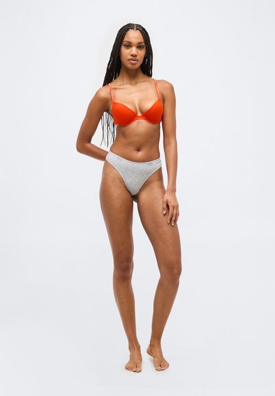 Γυναίκα με μακριά πλεξούδες που φοράει πορτοκαλί σουτιέν και γκρι εσώρουχα Emporio Armani, στέκεται ξυπόλητη μπροστά σε ένα απλό φωτεινό φόντο.