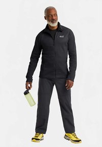 Giacca in pile nera con zip, pantaloni abbinati e scarpe da trekking gialle. Tiene in mano una bottiglia d'acqua verde chiaro. Tessuto morbido, vestibilità atletica, design funzionale.