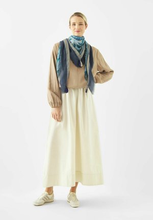 Donna in piedi che indossa una camicetta beige, una gonna lunga color crema, sneakers bianche e una sciarpa blu con motivo attorno al collo e alle spalle.