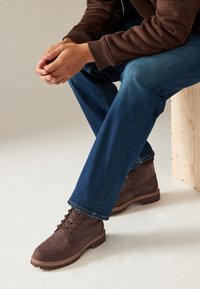 Bottes courtes en cuir marron avec des lacets superposés et des semelles robustes, assorties à un jean en denim bleu, posées sur un tabouret en bois devant un fond neutre.