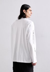 Garās piedurknes baltā t-kreklā ar taisnu griezumu, ar melnām grafiskām detaļām gar vienu piedurknu un mīkstu kokvilnas tekstūru.