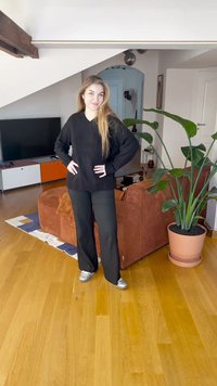 Črn prevelik pulover, črne hlače in srebrni športni copati. Notranja postavitev z rjavim kordurovim kavčem in zeleno rastlino v lončku. Lesena tla.