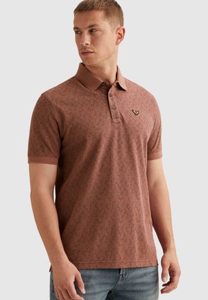 Mężczyzna w brązowej, wzorzystej koszulce polo z krótkim rękawem, kołnierzykiem i guzikami, w jasnoniebieskich dżinsach, stojący na jednolitym tle.