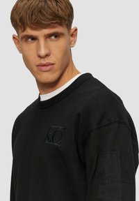 Zwarte sweater met een ronde hals, voorzien van een textuur logo patch. De stof lijkt dik; de linkermouw heeft een pocketdetail.