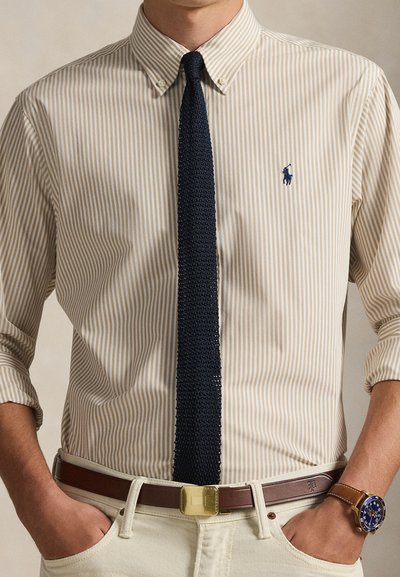 Μπεζ και κρεμ ριγέ πουκάμισο με γιακά με κουμπιά, σετ με υφασματοποιημένη ναυτική γραβάτα, καφέ δερμάτινη ζώνη και ανοιχτόχρωμο παντελόνι.