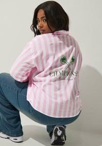 Rosa och vita randiga skjorta med knappar och en broderad grön grafik av palmträd och tennisracketar på baksidan, matchad med blå jeans.