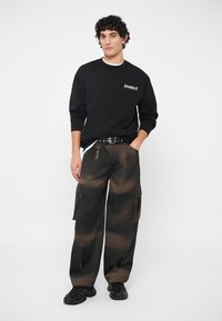 Μαύρη μπλούζα με κουκούλα που γράφει "Double" σε λευκό χρώμα, φορεμένη με καφέ και μαύρο cargo παντελόνι με πλαϊνές τσέπες και άνετη εφαρμογή. Μαύρα αθλητικά παπούτσια.