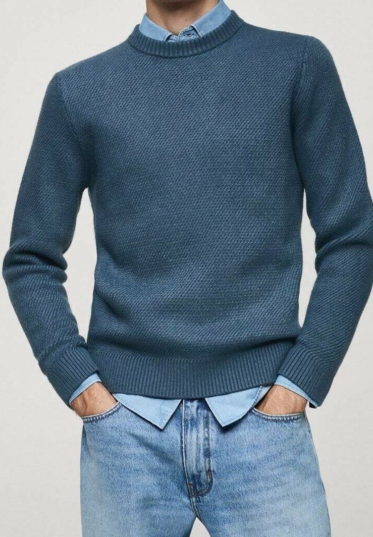 Homme portant un pull en maille bleu sur une chemise bleu clair boutonnée, les mains dans les poches d'un jean en denim bleu clair.