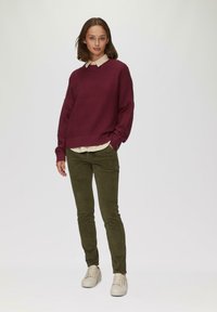 Pull à côtes bordeaux sur une chemise à col crème, associé à un pantalon slim-fit vert olive et des baskets beiges, sur un fond neutre.