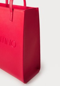 Κόκκινη τσάντα tote από ψεύτικο δέρμα με δομημένο σχήμα, που διαθέτει ανάγλυφο λογότυπο της μάρκας, δύο χερούλια, και λεία υφή.