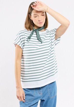 Femme portant une chemise blanche à rayures vertes et un pantalon en jean, avec un foulard vert autour du cou, le bras droit levé, les yeux fermés dans une pose détendue.