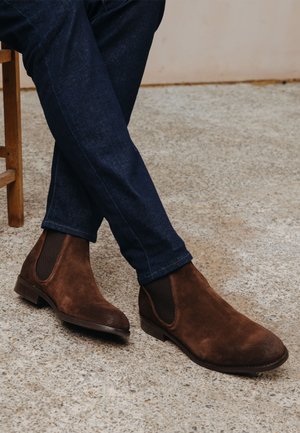 Brown suede Chelsea boots with elastic side panels, flat soles, rounded toes, worn with dark blue denim jeans, set on a textured surface.