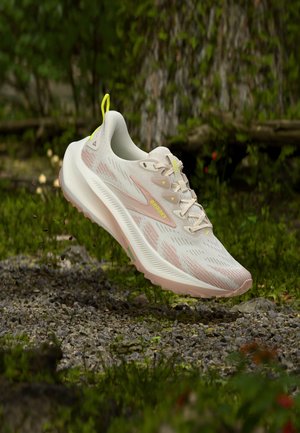 Lyse beige Brooks løbesko med pink accenter og gul trækflap, ophængt over grusstien med grøn bevoksning i baggrunden.
