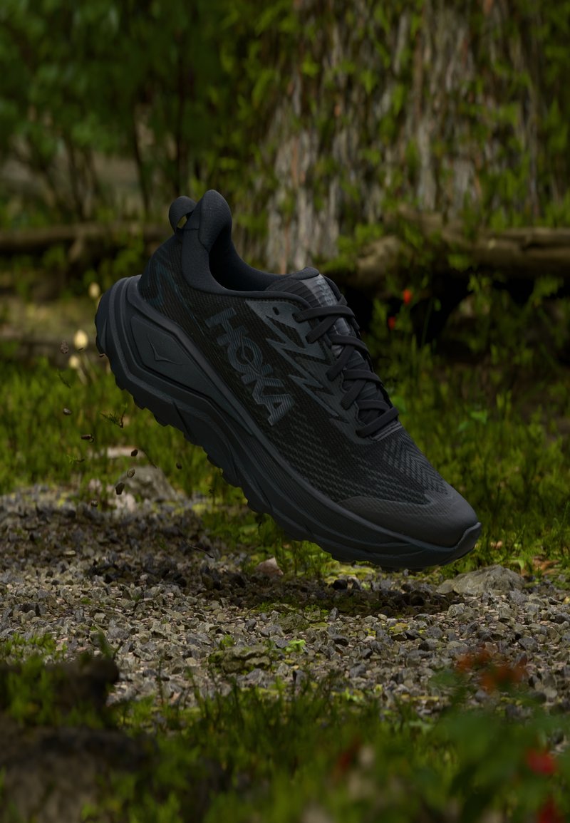 Μαύρο αθλητικό παπούτσι Hoka στον αέρα πάνω από μονοπάτι με χαλίκι, με πράσινη βλάστηση και κορμό δέντρου στο φόντο.
