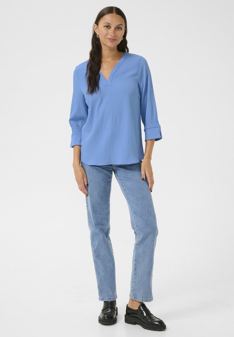 Blouse bleu clair à manches longues avec un col en V et un tissu texturé, assortie à un jean droit délavé et des mocassins noirs épais.