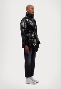 Chaqueta acolchada negra brillante con un alto cuello y cinturón, con un diseño acolchado. Complementada con jeans oscuros y zapatos plateados.