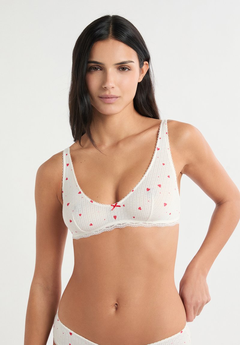 Weißer gerippter BH mit roten Herzmustern, ausgestattet mit einem Spitzenbesatz und einer kleinen roten Schleife in der Mitte. Weicher Stoff mit verstellbaren Trägern.