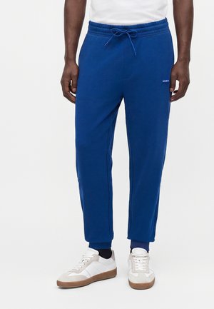 Uomo con pantaloni della tuta blu con coulisse e sneakers bianche, in piedi davanti a uno sfondo semplice.