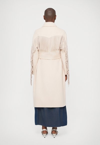 Beige ullkappa med fransdetaljer på baksidan, krage och midjebälte, i kombination med en lång denimkjol och klackskor.