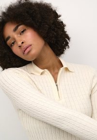 Top a maglia color crema con colletto con zip e motivo testurizzato, caratterizzato da un design aderente e maniche lunghe.
