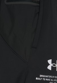 Schwarzes Under Armour Kleidungsstück mit einer Reißverschlusstasche und weißem Logo über dem Text "Ursprünge der Leistung."