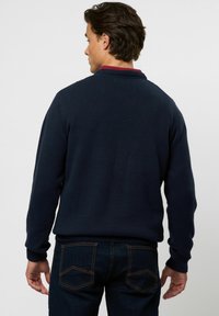 Mann von hinten, trägt einen dunkelblauen Pullover über einem roten Hemd und dunkelblaue Jeans mit oranger Naht auf den Gesäßtaschen.