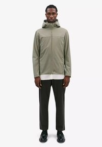 Olijfgroene softshell jack met een volledige rits, verstelbare capuchon en dubbele voorzakken. Gecombineerd met zwarte broek en zwarte schoenen.