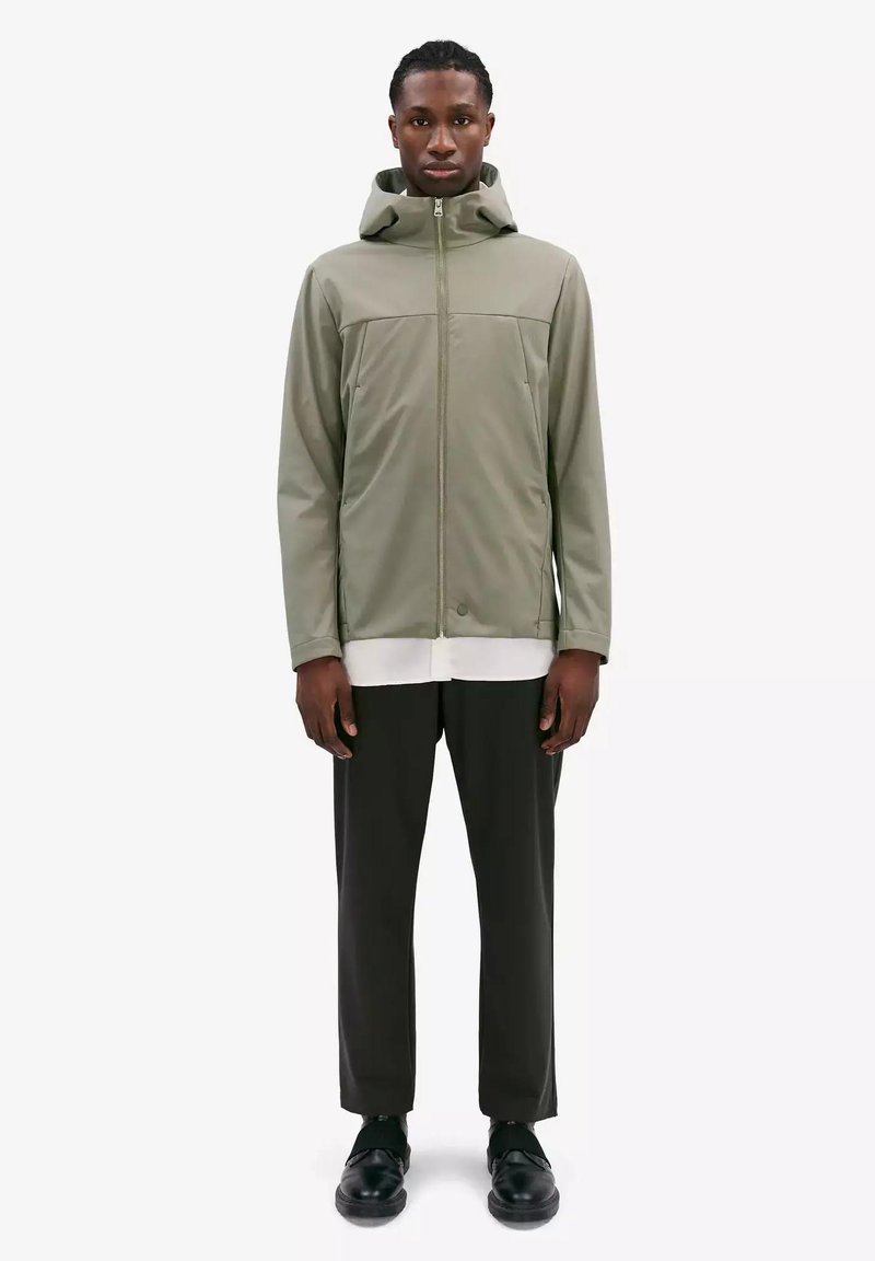 Olijfgroene softshell jack met een volledige rits, verstelbare capuchon en dubbele voorzakken. Gecombineerd met zwarte broek en zwarte schoenen.