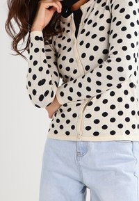 Cárdigan beige con lunares negros, botón delantero, puños de canalé y textura suave, combinado con jeans azul claros.