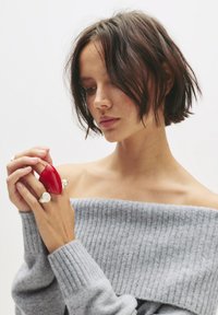 Piros gyűrű organikus formával, ezüst díszítéssel ellátott gyűrűk mellett viselve. Szürke bordázott, off-shoulder pulóver. Simá, minimalista dizájn.