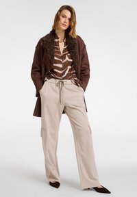Veste marron avec un col texturé, blouse à motifs marron et crème, et pantalon cargo beige clair. Des ballerines marron complètent l'ensemble.