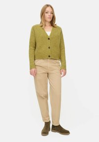 Groene wollen cardigan met een V-hals en knoopsluiting, gecombineerd met beige broek en donkergroene enkellaarsjes, tegen een effen achtergrond.