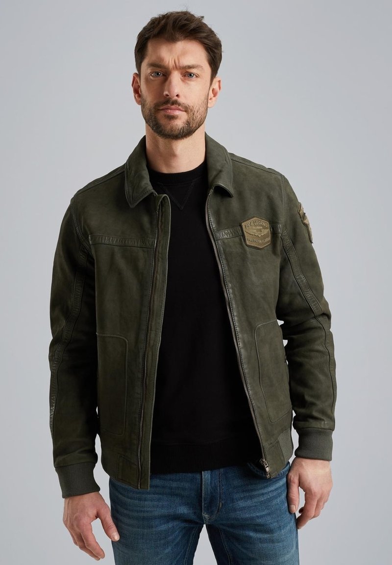 Chaqueta de cuero verde oliva con cremallera en la parte delantera, bolsillos laterales, parches en los hombros y puños acanalados. Lleva encima un suéter de punto negro.