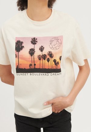 Person iført en hvid T-shirt med et tryk af solnedgang og palmer og teksten "SUNSET BOULEVARD DREAMS" foran.