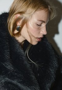 Musta tekoturkistakki, jossa on kiiltävä pinta. Kuvattavalla on vaaleanblondit hiukset ja hänellä on ylisuurikokoinen musta korvakoru. Luonnonvalo korostaa yksityiskohtia.