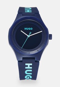 Reloj de goma azul marino con acentos azules, esfera redonda y manecillas de horas y minutos blancas. Presenta el texto "HUGO" en la correa y una banda texturizada.
