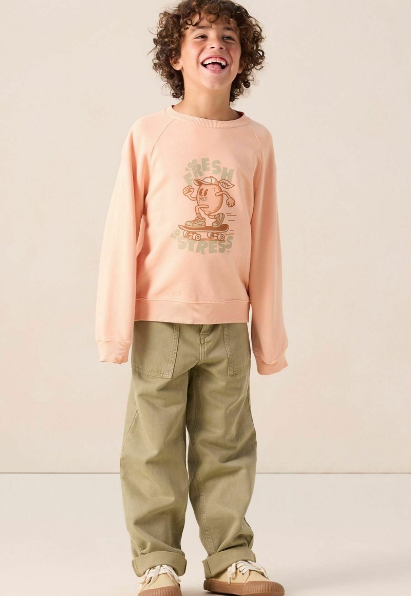 Un sweat-shirt rose clair avec un graphisme d'un personnage en train de faire du skate, associé à un pantalon ample de couleur olive. Des chaussures marron clair complètent la tenue.