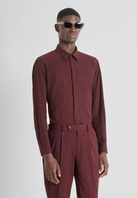 Camisa de manga larga burdeos con cuello, combinada con pantalones a juego que presentan un cinturón y detalles de botones. Gafas de sol negras completan el look.