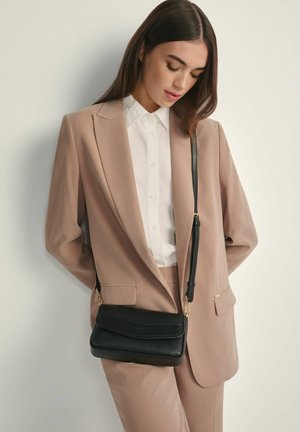 Kobieta w beżowym garniturze i białej koszuli stoi na tle białej ściany, nosząc czarną skórzaną torebkę na ramię z złotymi okuciami.