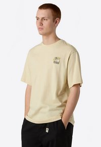 Hellbeiges Baumwoll-T-Shirt mit kurzen Ärmeln, Rundhalsausschnitt und kleinem Grafikdruck auf der linken Brust mit Blumen und industriellen Elementen.