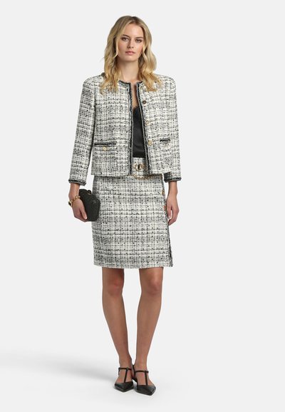 Kvinde iført sort og hvid tweedjakke og nederdel med gyldne knapper, holder en sort clutch og bærer sorte T-remme-sandaler.