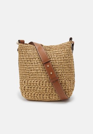 Gevlochten strooien bucketbag met brede bruine verstelbare leren schouderriem en gouden metalen beslag op een witte achtergrond.