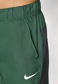 Zaļi sporta šorti, kas izgatavoti no elpojoša auduma, ar elastīgu jostasvietu, sānu kabatām un melniem akcentiem, ar balto Nike logotipu.