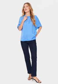 Blusa blu a maniche corte con scollatura arricciata, abbinata a jeans in denim scuro e sandali neri con cinturini. La blusa presenta un motivo testurizzato.
