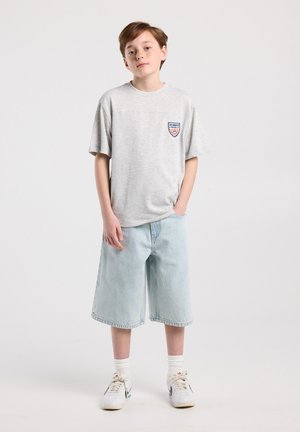 Boy with short brown hair wearing a gray Los Angeles T-shirt, light blue denim shorts, white socks, and white sneakers standing against a plain backdrop.